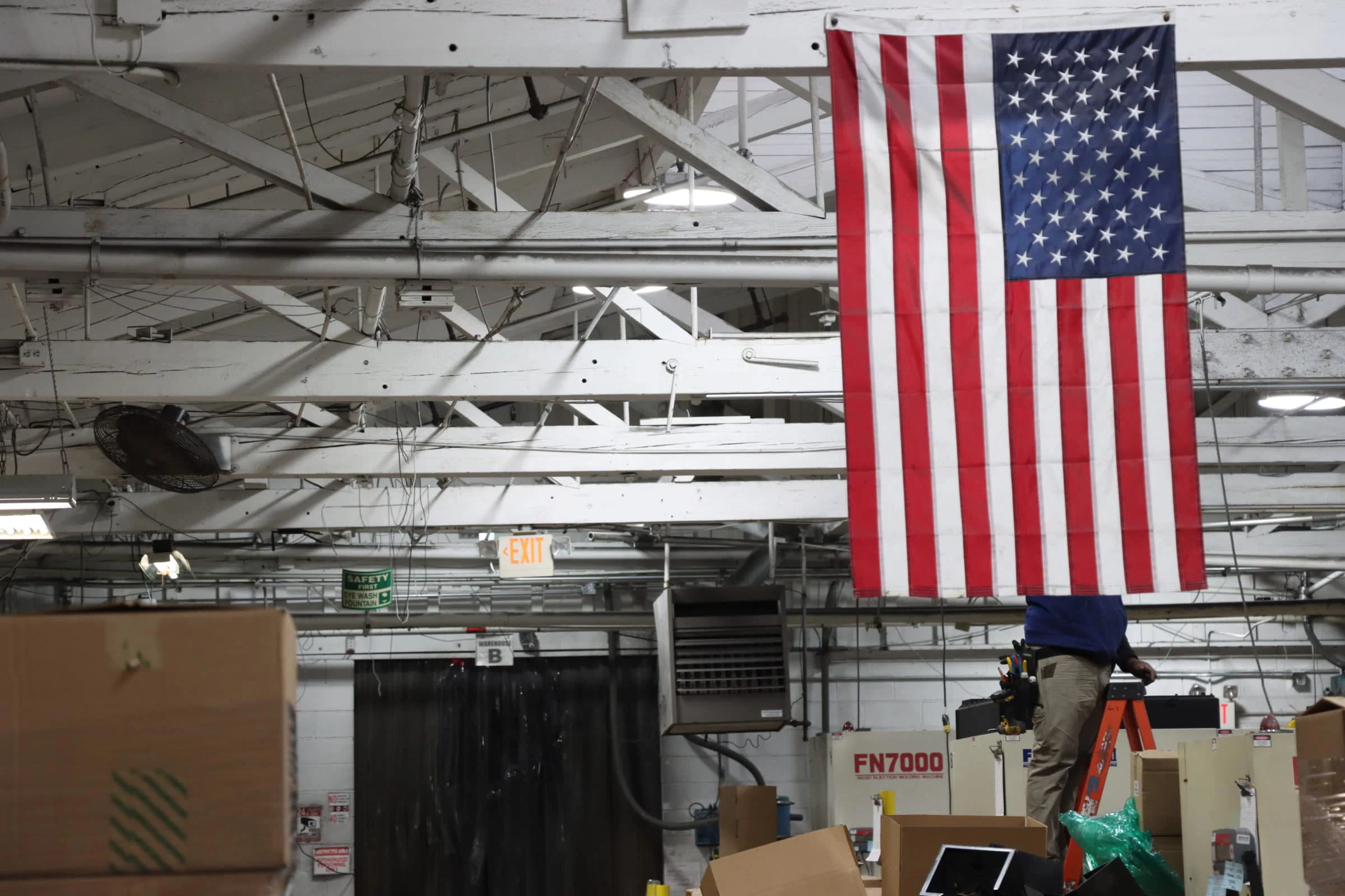 IMG_5100 A man repairs a light on a beam behind an American flag that hangs over a factory floor.