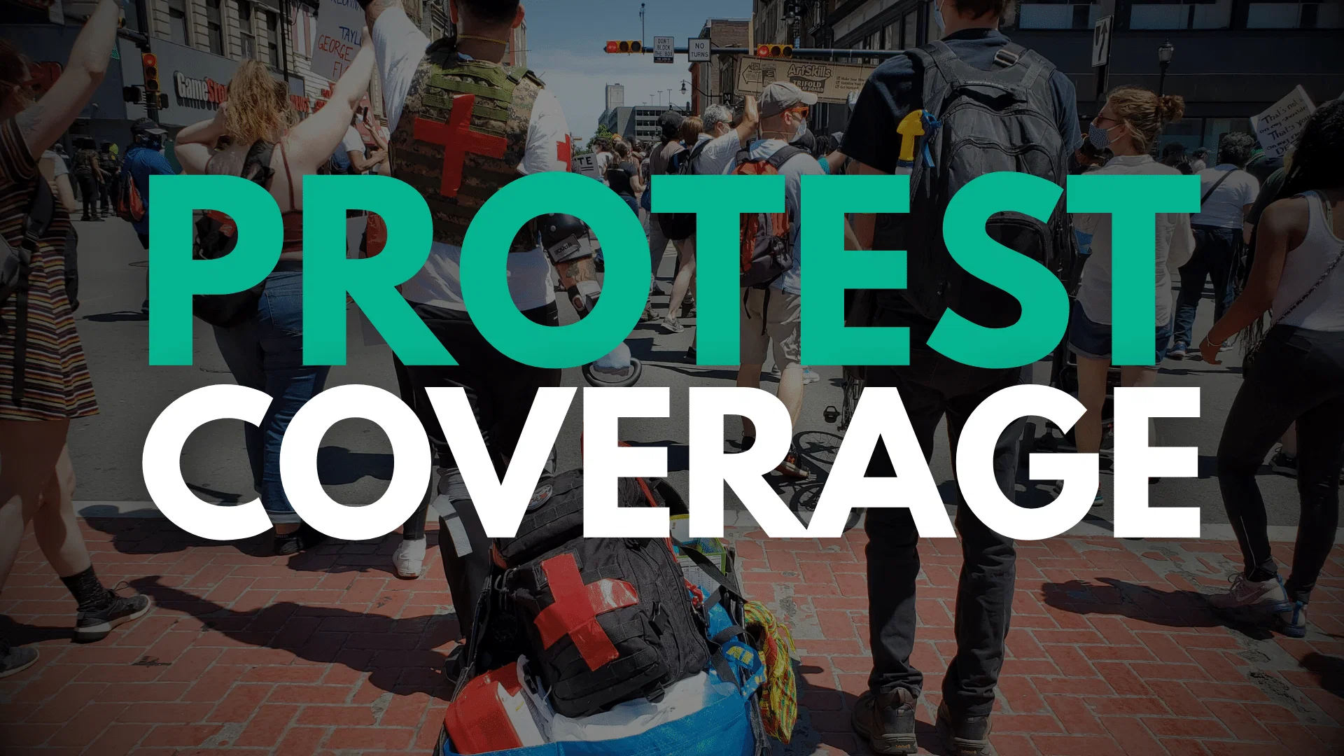 protest coverage feat Protesters gather on a city street carrying signs and banners during a demonstration. Some participants kneel on the brick crosswalk while others stand nearby, with urban buildings and traffic signals visible in the background.